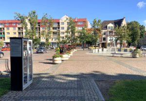 Widok na część ulicy. Pierwszy plan szafa bookcrossingowa, niedaleko zegar słoneczny a w tle parking, domy i sklepy.