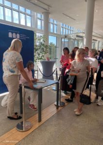 Kobieta podpisuje książki, obok stolika stoi kobieta w jasnych spodniach z drugiej strony kolejka ustawiona po autograf.