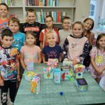 Dzieci stoją na tle regałów z książkami i prezentują swoje prace.
