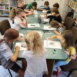 Dzieci siedzą przy stole i kolorują kolorowanki. W tle widać regały z książkami.
