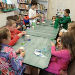 Dzieci siedzą przy stole i robią instrumenty z materiałów z recyklingu. W tle widać regały z książkami.