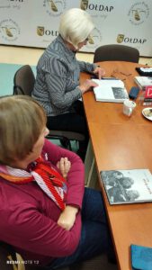 Dwie kobiety siedzące za stołem, jedna ma otwartą książkę i czyta.