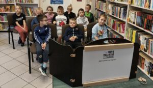 Grupa dzieci ogląda teatrzyk kamishibai. W tle widać regały z książkami.