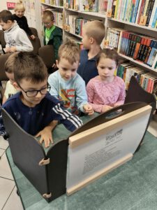 Grupa dzieci ogląda teatrzyk kamishibai. W tle widać regały z książkami.