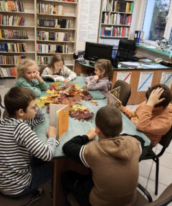 Dzieci siedzą przy stole na którym leżą książki i jesienne liście w tle widać regały z książkami.