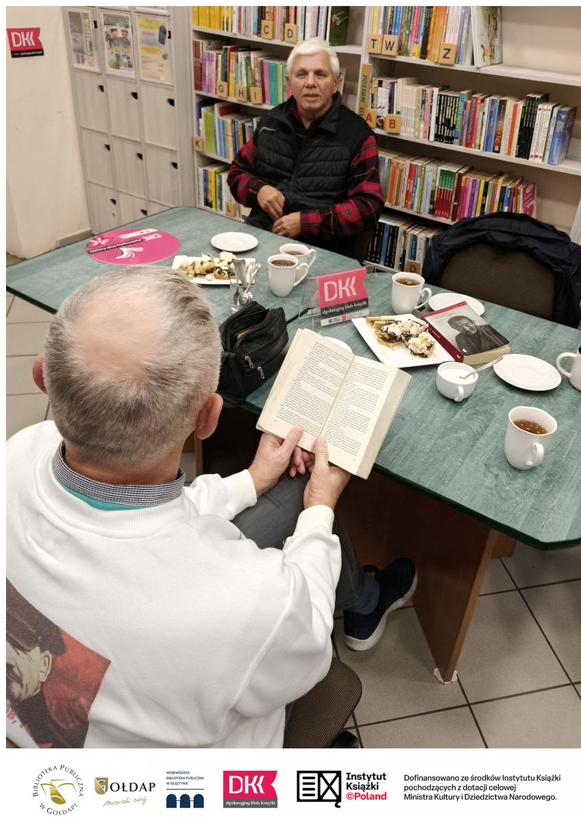 Dwie osoby siedzą przy stoliku na którym znajduje się ciasto i kubki z kawą i herbata. W tle widać regały z książkami