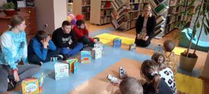 Grupa dzieci i młodzieży siedzi na podłodze w bibliotece, słuchając kobiety czytającej książkę. Wokół nich leżą książki i edukacyjne.