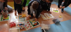 Uczniowie siedzą na podłodze, budując modele z klocków konstrukcyjnych (LEGO) zgodnie z instrukcjami obrazkowymi, używając posortowanych klocków z plastikowych pojemników.