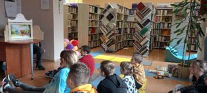 Dzieci siedzą na piankach w bibliotece oglądając mały, drewniany teatrzyk kamishibai stojący na stole. W tle widać regały z książkami.