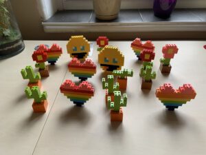 Małe konstrukcje zbudowane z klocków LEGO w tym kaktusy, kwiaty, tęczowe serca i figurki emoji, ułożone na jasnym stole.