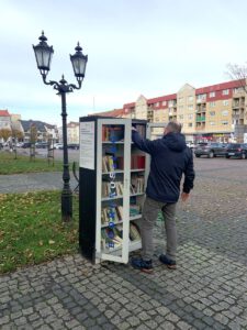 Mężczyzna w ciemnej kurtce i spodniach sięga do otwartej szafki bookcrossingowej, przeglądając książki. Stoi na bruku w parku miejskim, w tle widać budynki.