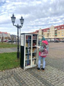 Kobieta w różowej bluzie, dżinsach i czapce (równie kolorowej) stoi przy otwartej szafce bookcrossingowej i czyta książkę. W tle miejski krajobraz.