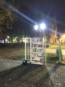 Oświetlona witryna z książkami (biblioteczka bookcrossingowa) na zewnątrz w nocy, pod stylową latarnią parkową. Czekająca na czytelników szafka rzuca długi cień na brukowaną ścieżkę.