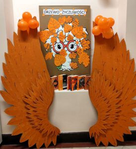 Skrzydła pomarańczowe ustawione pod ścianą pomiędzy nimi stoją książki oraz wisi tablica z drzewem życzliwości.