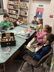 Dzieci siedzą przy stole na którym leżą książki i jesienne liście, szyszki i kasztany.