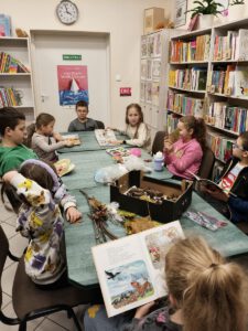 Dzieci siedzą przy stole na którym leżą książki i jesienne liście w tle widać regały z książkami.