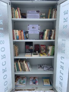 Otwarta metalowa szafka biblioteczna typu bookcrossing, wypełniona książkami i czasopismami, stojąca na zewnątrz w zimowej scenerii. Na półkach widoczne są polskie wydania książek oraz plakaty akcji „Tata też czyta” i „Radość czytania”.