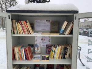 Zbliżenie na przeszkloną szafkę do bookcrossingu pokrytą warstwą śniegu. Wewnątrz, na białych półkach, równo ułożone rzędy książek oraz ogłoszenia Biblioteki Publicznej w Gołdapi dotyczące akcji „Tata też czyta” i „Radość czytania”.