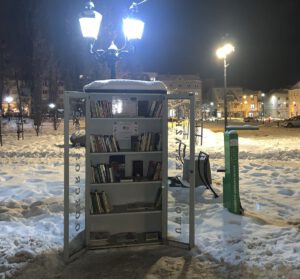 Otwarta, oświetlona latarnią szafka do bookcrossingu nocą w zaśnieżonym parku. Ciepłe światło lampy pada na półki z książkami i biały napis na szklanych drzwiach. W tle widać oświetlone budynki i ulice miasta w zimowej scenerii.