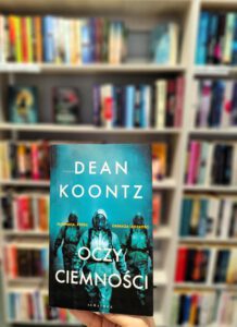 Na pierwszym planie książka trzymana w ręku, w tle regał z książkami.