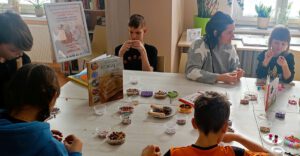 Zdjęcie przedstawia osoby siedzące nawlekające koraliki na żyłkę.