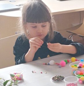 Fotka przedstawia dziewczynkę nawlekającą koralika na żyłkę.