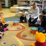 Dzieci siedzące na dywanie w kółeczku, kobieta czyta książkę.