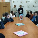 Zdjęcie przedstawia dzieci siedzących przy stole, stojącą Pania Dyrektor wraz z funkcjonariuszem Policji.