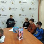 Na zdjęciu widoczne dzieci siedzące przy stole i pani Policjant czytająca fragment książki.