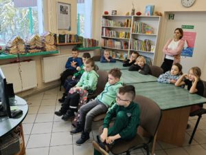 Grupa dzieci siedzi na krzesłach i słucha czytanego tekstu i ogląda . W tle widać regały z książkami.
