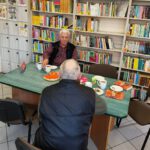 1. Dwie osoby siedzą przy stoliku na którym znajduje się ciasto i kubki z kawą i herbata. W tle widać regały z książkami.