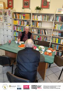 1. Dwie osoby siedzą przy stoliku na którym znajduje się ciasto i kubki z kawą i herbata. W tle widać regały z książkami.