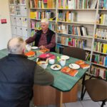 2. Dwie osoby siedzą przy stoliku na którym znajduje się ciasto i kubki z kawą i herbata. W tle widać regały z książkami