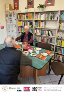 2. Dwie osoby siedzą przy stoliku na którym znajduje się ciasto i kubki z kawą i herbata. W tle widać regały z książkami