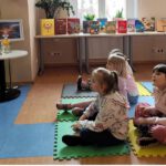 Dzieci siedzące na matach przed nimi siedzi starsza pani.