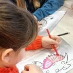 Dziewczynka na zdjęciu koloruje kredką dinozaura na różowo.