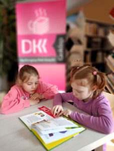 Na zdjęciu widoczne dwie dziewczynki, jedna z nich czyta książkę.