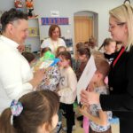 Grupa dzieci wokół mężczyzny i kobiety, która wręcza biały skoroszyt.