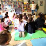 Grupa dzieci siedząca tyłem na piankach przed nimi stoi pan w białym golfie.