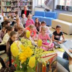 Pan siedzący na fotelu z książką, przed nim siedzi grupa dzieci i dwie kobiety.