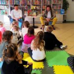 Dzieci siedzące tyłem, a przed nimi siedzi pan w fotelu z książką w ręku.