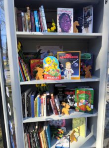 Szafa bookcrossingowa wypełniona książkami, dodatkowo ustawione są króliczki drewniane i tulipany w kolorze żółtym.