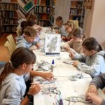 Zdjęcie przedstawia dzieci siedzących i wykonujących pracę przy długim stole na którym stoi ramka z wykonaną pracą.