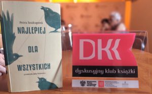 Książka ze stojakiem na którym widniej napis Dyskusyjny Klub Książki w tle widać uczestniczki